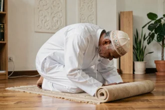 Homme musulman en djellaba blanche en position de prière sur un tapis de prière dans un appartement à Nantes
